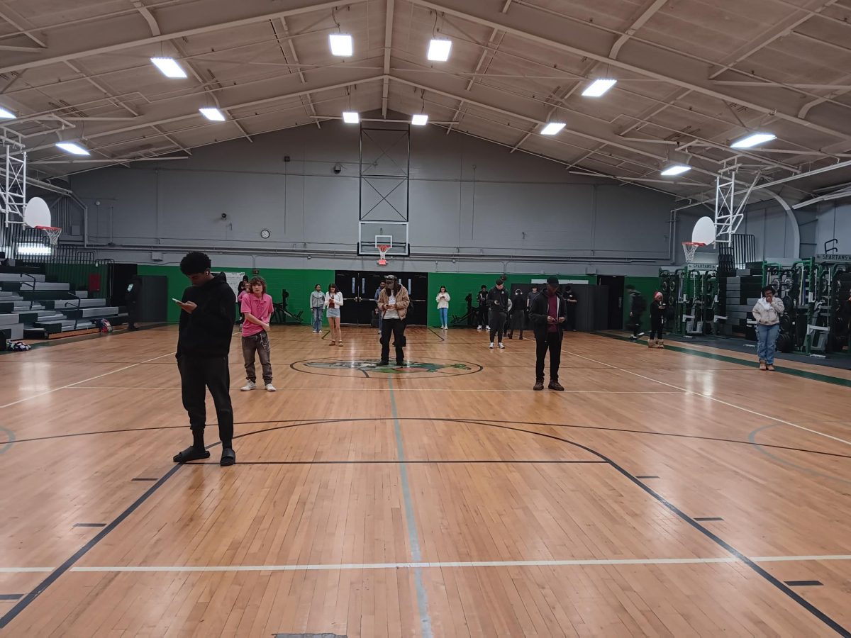Students stand on the gym floor, looking at their phones. Markings on the floor were used as alternatives to assigned seats for taking attendance.